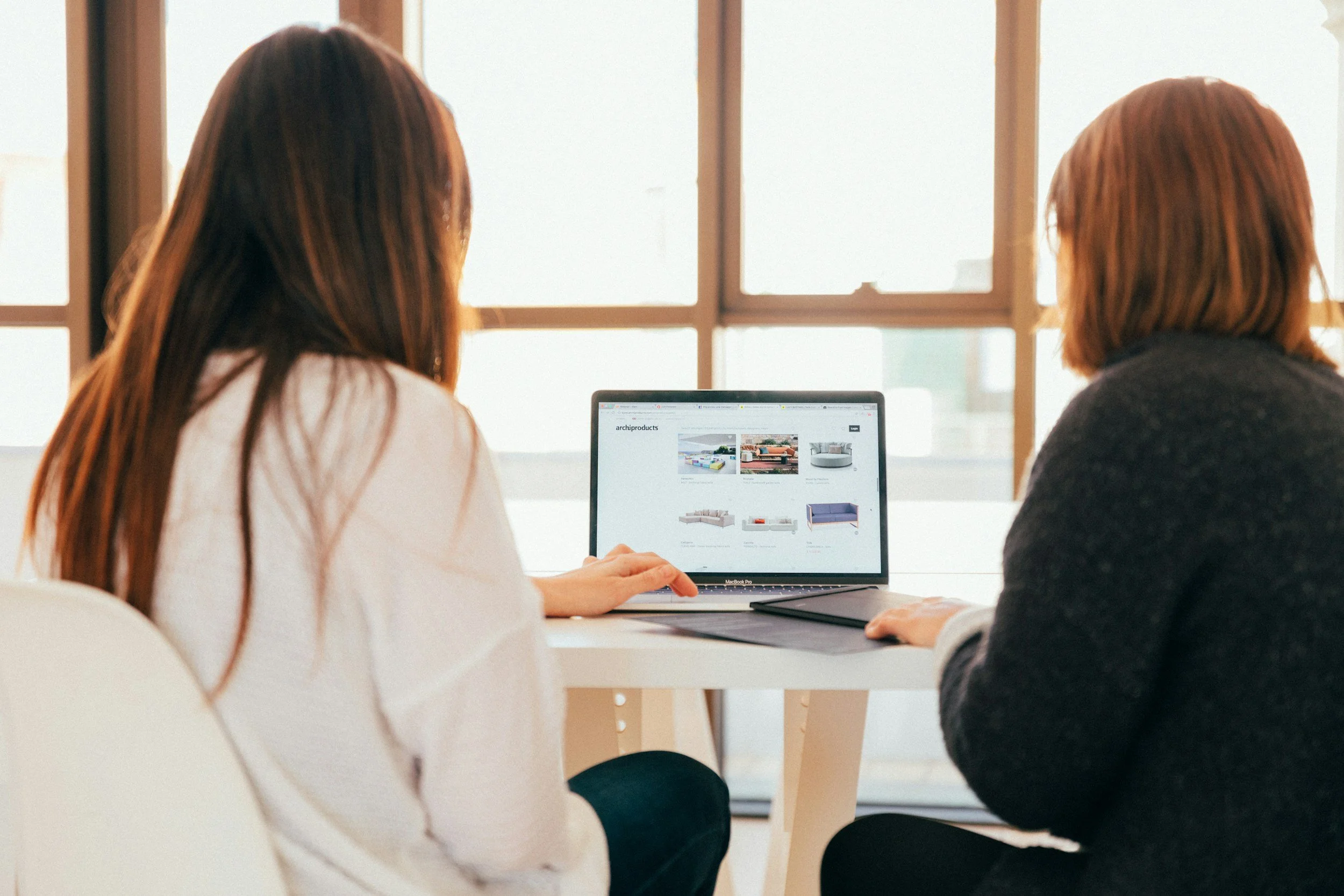 Two women sitting at a desk looking at a laptop showing furniture images on the screen.