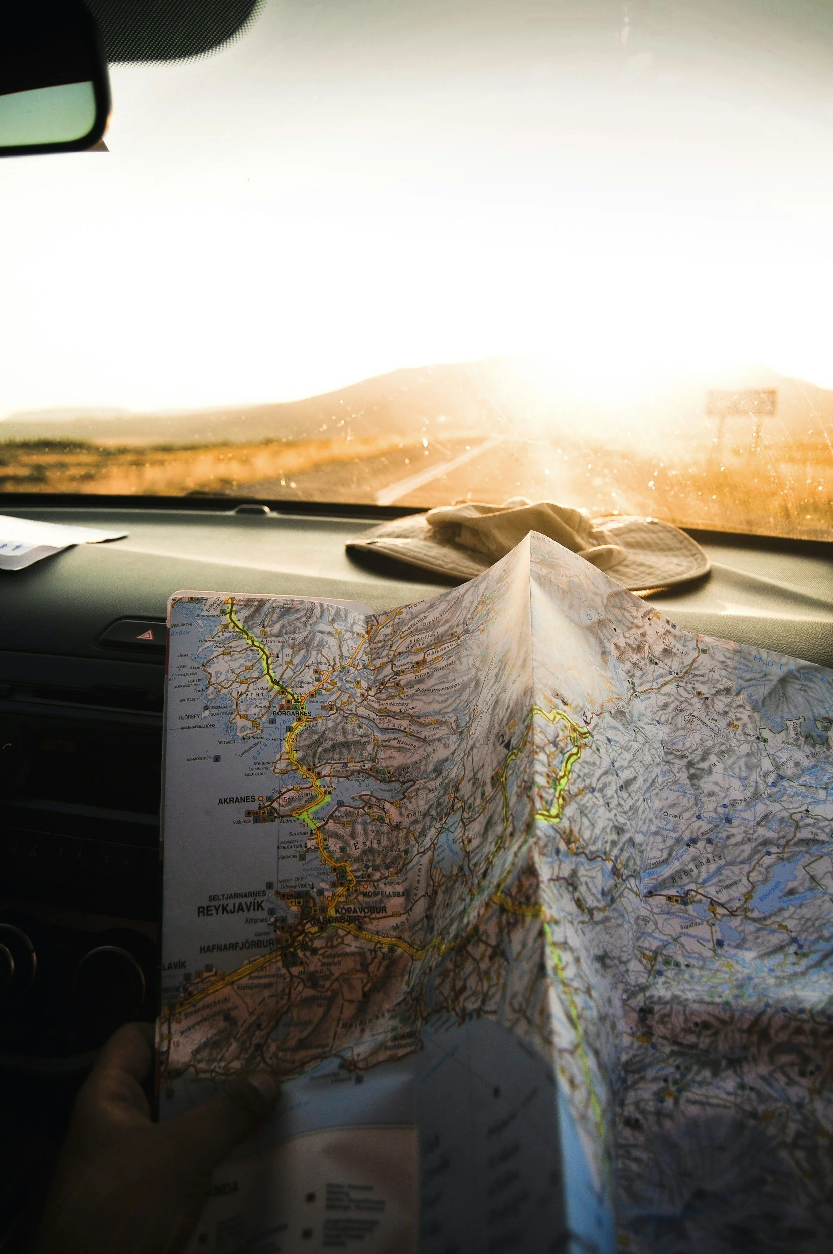 A person holding a paper map inside a vehicle, with a view of a road and sunset through the windshield.